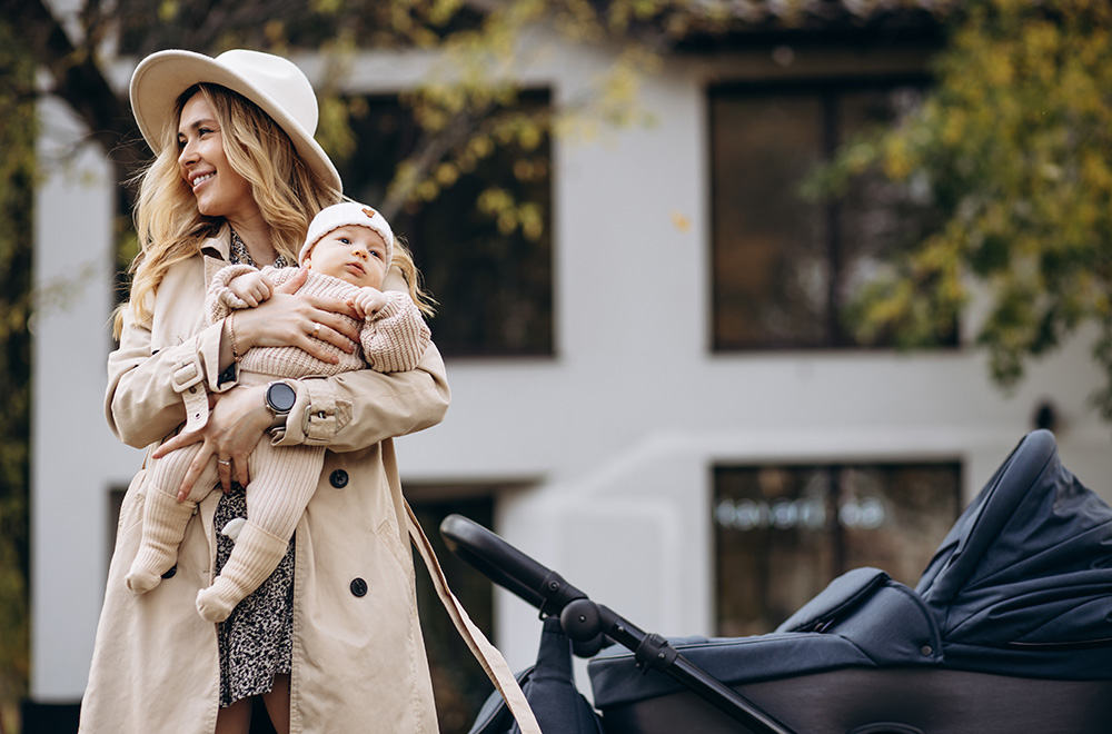 Žena drží v náručí bábätko a kráča vedľa kočíka pred budovou